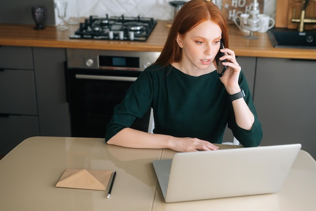 Blick aus der Vogelperspektive auf eine wütende Freiberuflerin, die mit dem Handy telefoniert und einen Laptop benutzt, der am Tisch in einer Küche mit modernem Interieur sitzt. Verärgerte junge Frau, die mit dem Support-Manager auf dem Handy spricht.