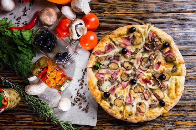 Blick aus der Vogelperspektive auf leckere Pizza mit Schinken, Zwiebeln, Oliven auf Holztisch, in der Nähe von Tomaten, Pilzen und anderem Gemüse