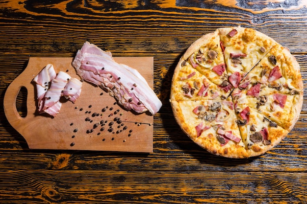 Blick aus der Vogelperspektive auf Pizza mit verschiedenen Belägen und Käse auf Holztisch, in der Nähe von Schneidebrett mit Speck und Gewürzen