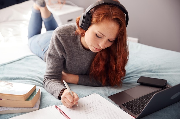 Blick nach unten auf Studentin mit Kopfhörern, die auf dem Bett liegt und am Laptop arbeitet