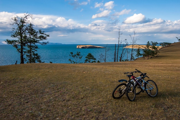 Blick über die Steppe auf die Insel im Baikalsee. Auf dem Feld stehen sie umarmt von zwei Fahrrädern. Am Rand der Küste wachsen Bäume, am Horizont des Berges. Wolken im Himmel.