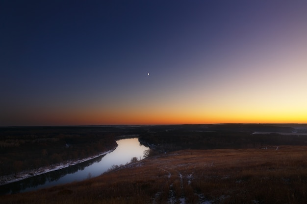 Blick vom Hügel zum Mond über den Fluss in der Abenddämmerung. Heller Abendhimmel nach Sonnenuntergang.