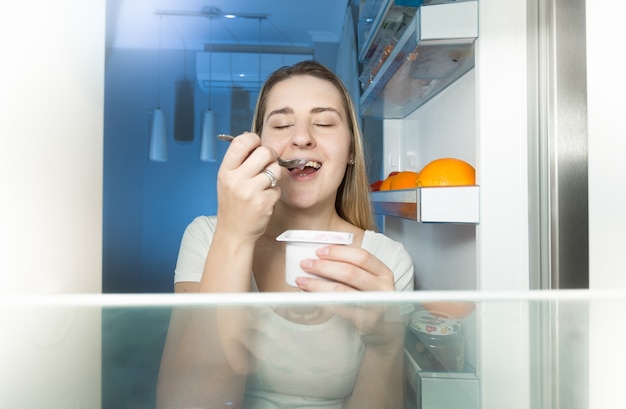 Blick vom Inneren des Kühlschranks auf eine junge Frau, die nachts Joghurt isst?