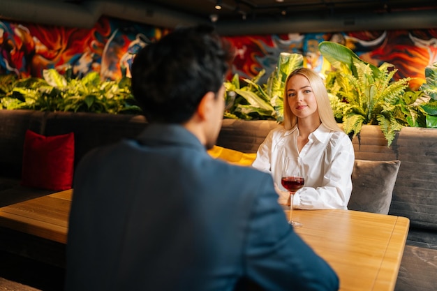 Blick von der Rückseite des nicht erkennbaren jungen Mannes auf die schöne blonde Frau, die mit einem Glas Rotwein am Tisch sitzt und ein festliches Abendessen im Restaurant hat