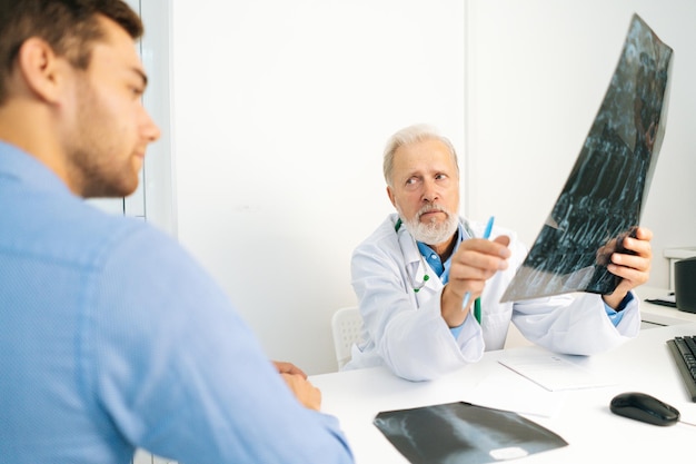 Blick von der Rückseite eines reifen männlichen Arztes, Radiologe, der die Ergebnisse der MRT-Untersuchung für einen jungen Patienten erklärt, der einen Schnappschuss mit Bildern zeigt, die eine MRT-Untersuchung im Klinikarzt analysieren, der die Testergebnisse überprüft
