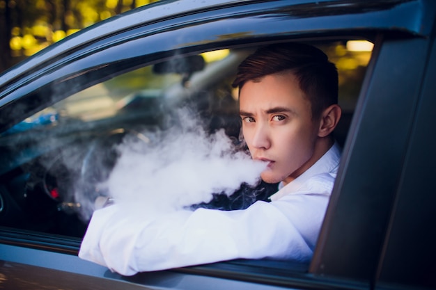 Blick von der Seite junger Mann, der eine E-Zigarette raucht, während er sein Auto auf einer städtischen Straße fährt. Autofahrer guckt aus dem Auto und raucht