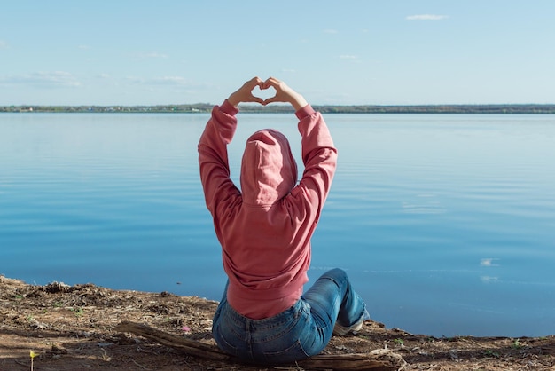 Blick von hinten auf eine Frau, die ihre Finger in Form eines Herzens gefaltet hat, während sie am See sitzt.