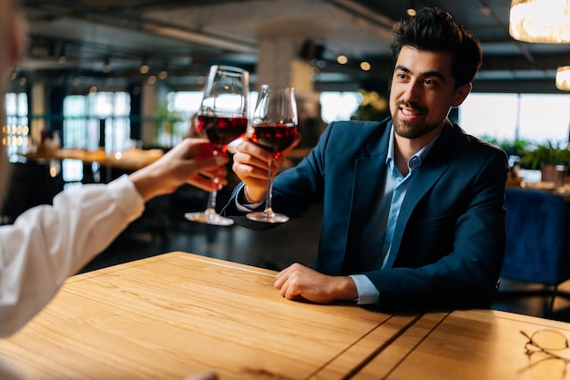 Blick von hinten auf eine unkenntlich klirrende Frau mit Rotweingläsern und einem lächelnden eleganten Mann im Anzug, der abends in einem schicken Restaurant am Tisch sitzt