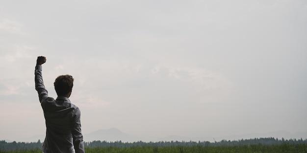 Blick von hinten auf einen erfolgreichen Geschäftsmann, der mit hoch erhobenem Arm in der Natur unter bewölktem Himmel steht