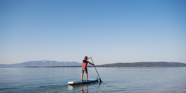 Blick von hinten auf einen jungen Mann, der auf einem SUP-Board paddelt, das auf ruhigem Meerwasser am Morgen schwimmt.