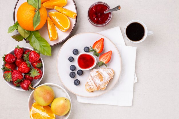 Blick von oben auf das Frühstück mit Tasse Kaffee, frischen Croissants und reifen Erdbeeren, Blaubeeren und saftigen Orangen auf weißem Tisch.