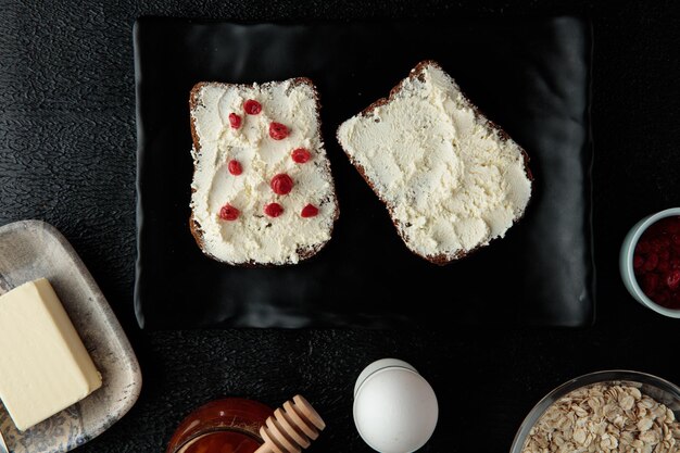 Blick von oben auf das Frühstücksset mit Hüttenkäse auf Brotscheiben mit roter Johannisbeere und Ei Hafer Quittenmarmelade Butter auf schwarzem Hintergrund