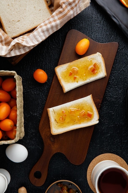 Blick von oben auf das Frühstücksset mit mit Marmelade bestrichenen Brotscheiben auf Schneidebrett und Orangenscheiben im Teller mit Kumquats Tasse Tee Ei auf schwarzem Hintergrund