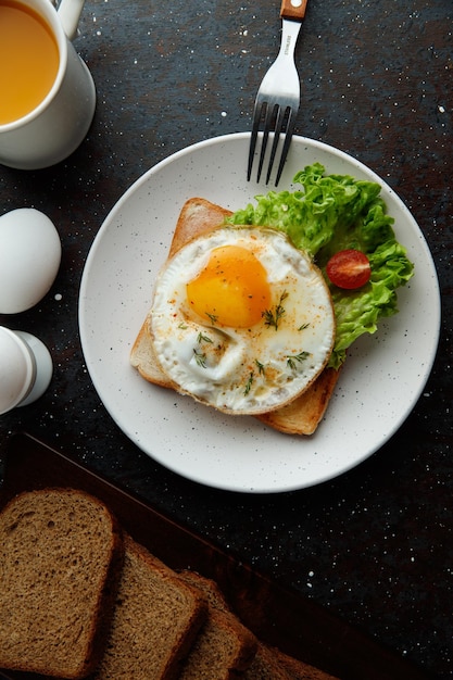 Blick von oben auf das Frühstücksset mit Spiegelei mit Salat und Tomate auf getrockneter Brotscheibe in Teller und Gabel mit Orangensafteiern und Brotscheiben auf schwarzem Hintergrund