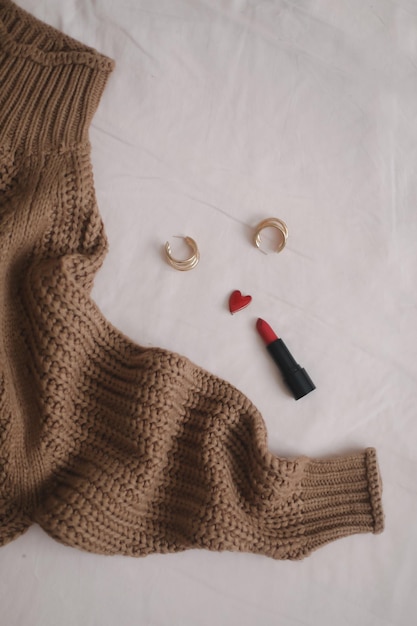 Blick von oben auf das lässige Outfit der Frau 39 mit warmem Strickpullover, rotem Lippenstift und Ohrringen auf beigem Hintergrund im Bett