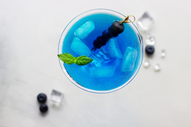 Blick von oben auf den blauen Cocktail im Martini-Glas mit Eiswürfeln, Minze und Blaubeeren