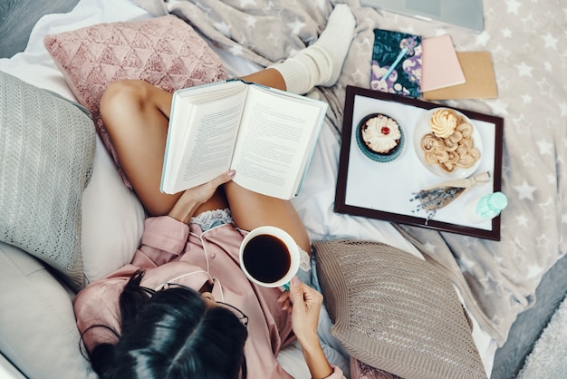 Blick von oben auf die schöne junge Frau im Pyjama, die den Morgenkaffee genießt, während sie zu Hause im Bett ruht