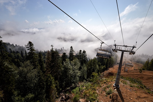 Blick von oben auf die Seilbahn mit mehreren Sitzen und gespannten Kabeln über Bäumen im tiefen Nebel. Berge des Nordkaukasus.