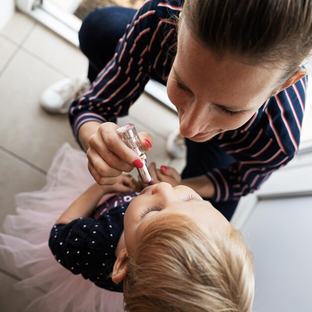 Blick von oben auf eine glückliche, modische junge Mutter, die ihrer Kleinkindtochter Lippenstift aufträgt.