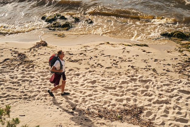 Blick von oben auf einen glücklichen reifen Mann, der an einem sonnigen Tag mit Rucksack am Meer entlang wandert?