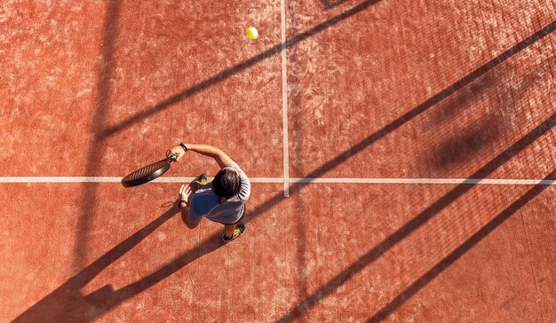 Blick von oben auf einen Paddle-Tennisspieler, der den Ball mit dem Schläger auf einem Außenplatz schlägt.