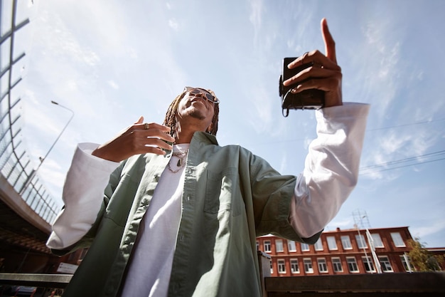 Blick von unten auf einen jungen aufgeregten afroamerikanischen Mann mit Walkman in der Hand, der gegen den blauen Himmel im städtischen Umfeld Hip-Hop spielt