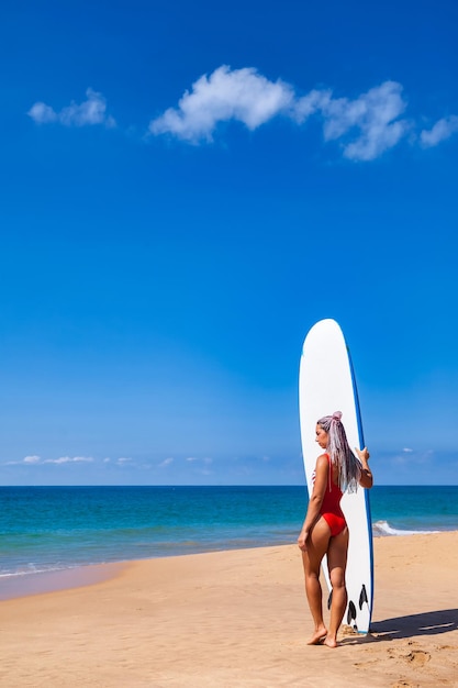 Blick zurück auf stilvolle junge Frau mit Surfbrett im Hintergrund blauer Himmel und Meer am Strand