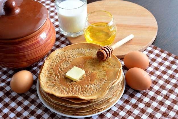 blini no prato com ovos, concha de mel, copo de leite e pote de cerâmica na toalha de mesa xadrez