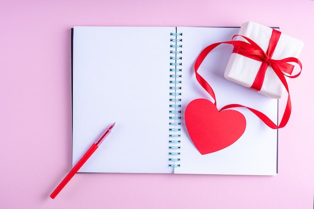 Bloc de notas abierto en blanco blanco, bolígrafo rojo, caja de regalo con cinta roja y forma de corazón de papel rosa