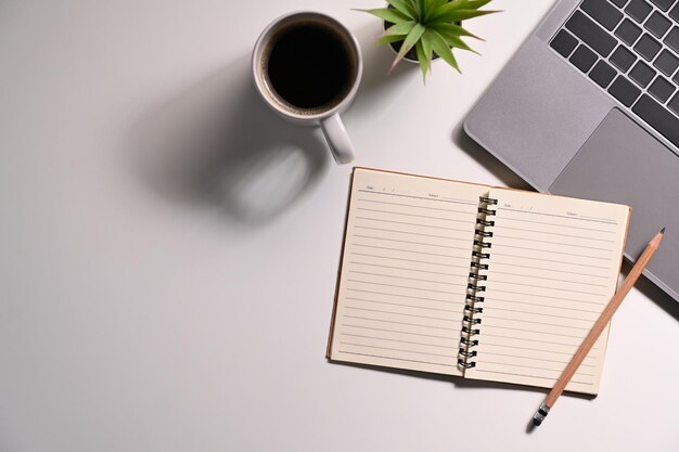 Bloc de notas en blanco, computadora portátil, taza de café y planta en maceta en el escritorio de oficina blanco