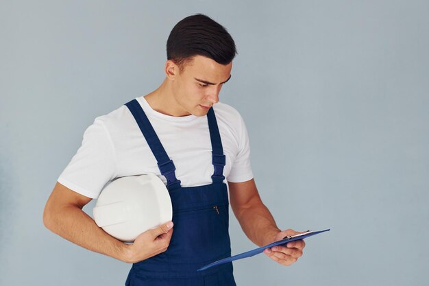 Foto con bloc de notas y casco trabajador de sexo masculino en uniforme azul de pie dentro del estudio contra el fondo blanco.