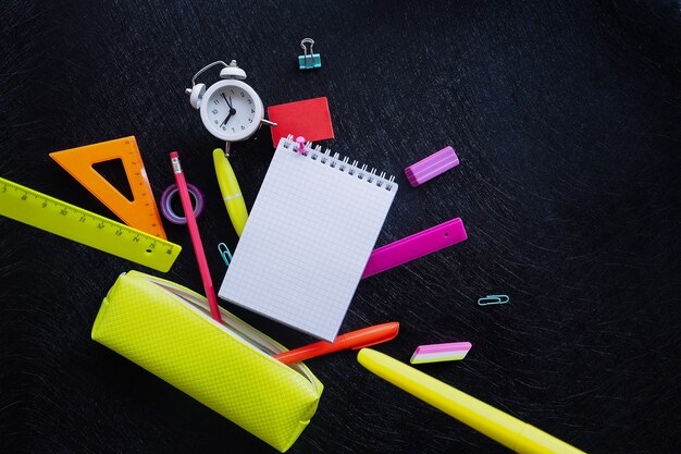 Bloc de notas a cuadros, reloj despertador y papelería multicolor cayendo de una caja de lápices de color amarillo brillante contra un fondo de textura negra.