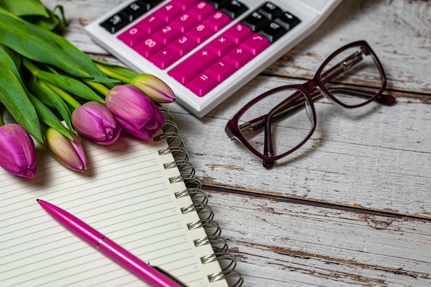 Bloc de notas de espacio de trabajo y bolígrafo esfera financiera calculadora aspecto festivo tulipanes y gafas de mujer en un primer plano de fondo de madera
