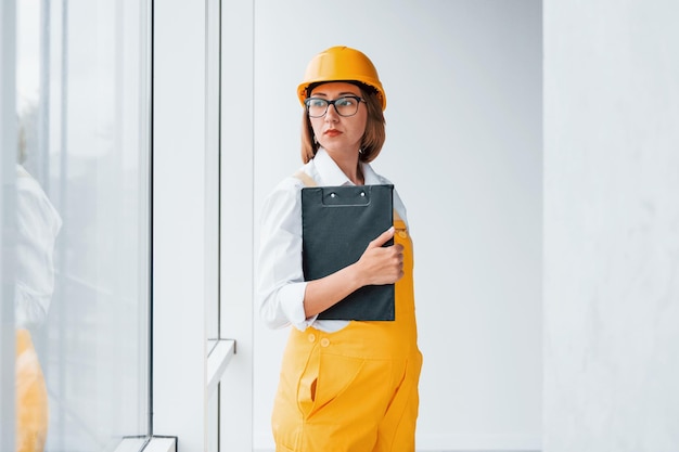 Con bloc de notas en las manos Trabajadora o ingeniera en uniforme amarillo y casco de pie en el interior