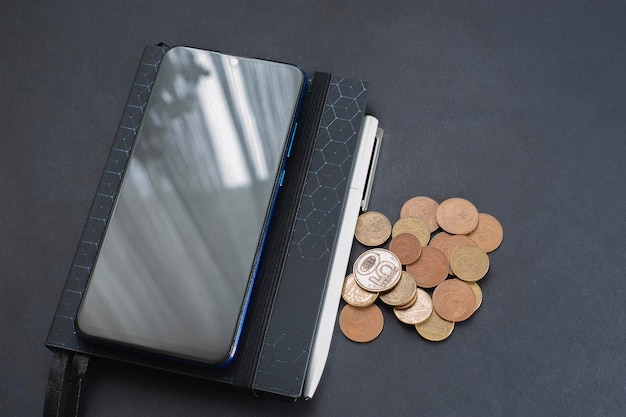 Bloc de notas y teléfono móvil con monedas en una mesa de fondo negro Cálculos financieros tonos negros