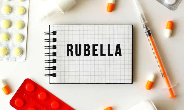 Bloc de notas con texto RUBELLA sobre una superficie blanca. Muy cerca se encuentran varios medicamentos.