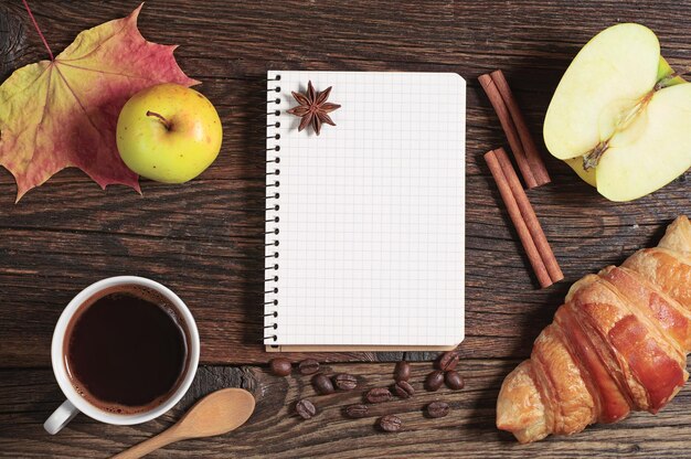 Bloco de notas aberto com página vazia, xícara de café quente e croissant na mesa de madeira escura, vista superior