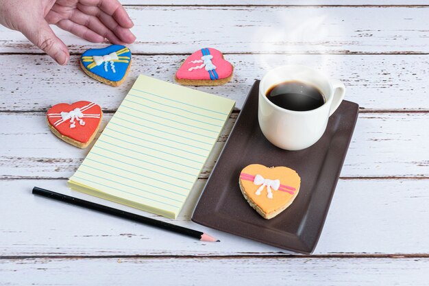 Bloco de notas ao lado de uma xícara de café, lápis e uma mulher pegando um biscoito.