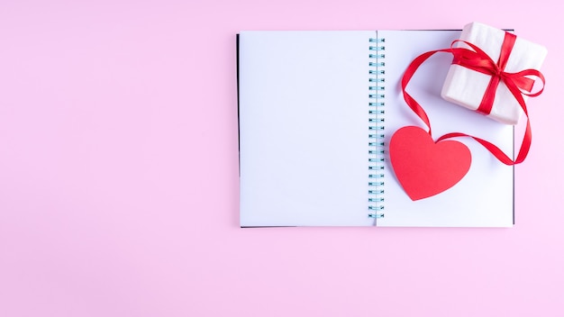 Bloco de notas branco em branco aberto, caixa de presente com fita vermelha e formato de coração de papel rosa em fundo rosa
