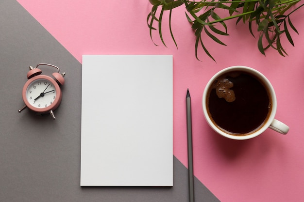 Bloco de notas, lápis, planta, xícara de café e despertador na mesa rosa e cinza