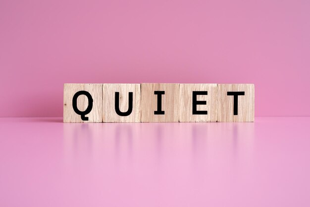 Blocos de madeira formam o texto QUIET contra um fundo rosa