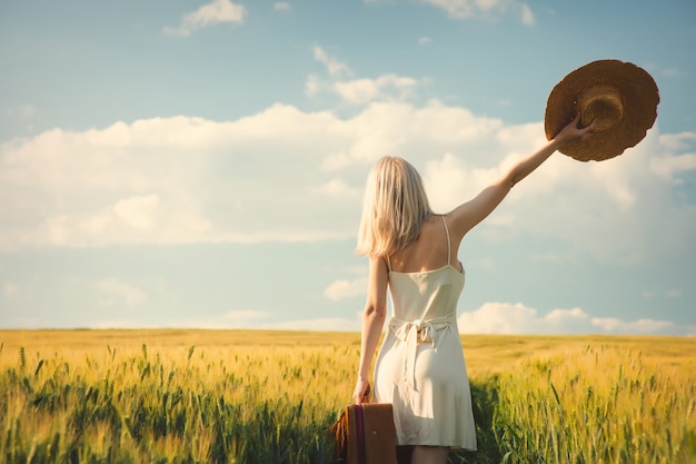 Blode mujer con sombrero con maleta en campo de trigo en la hora del atardecer