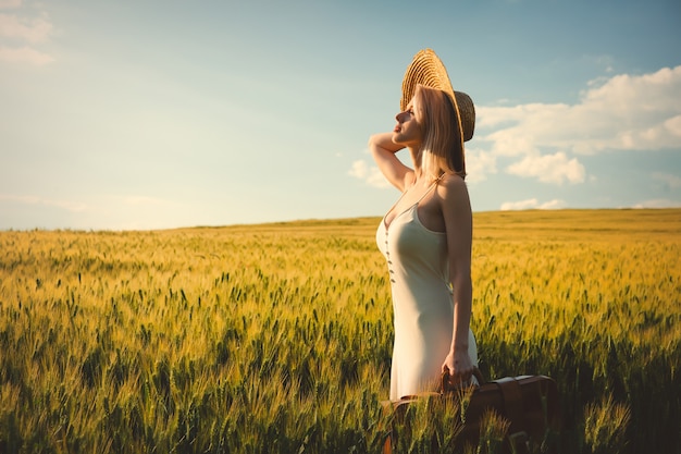 Blode mujer con sombrero con maleta en campo de trigo en la hora del atardecer