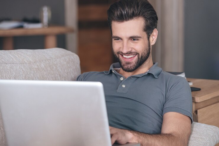 Foto blogger alegre y guapo. alegre joven guapo usando su computadora portátil con una sonrisa mientras está sentado en el sofá en casa