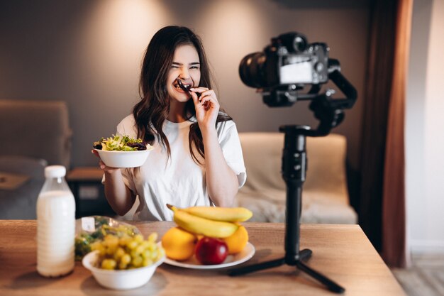 Blogger de alimentos saludables joven comiendo ensalada vegana fresca en el estudio de la cocina, tutorial de filmación en la cámara para el canal de video. La mujer influyente no muestra comida chatarra, habla de una alimentación saludable.