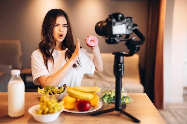 Blogger de alimentos saludables joven mujer cocinando ensalada vegana fresca de frutas y diciendo no a los dulces en el estudio de la cocina tutorial de filmación en la cámara para el canal de video Mujer influyente no muestra comida chatarra para