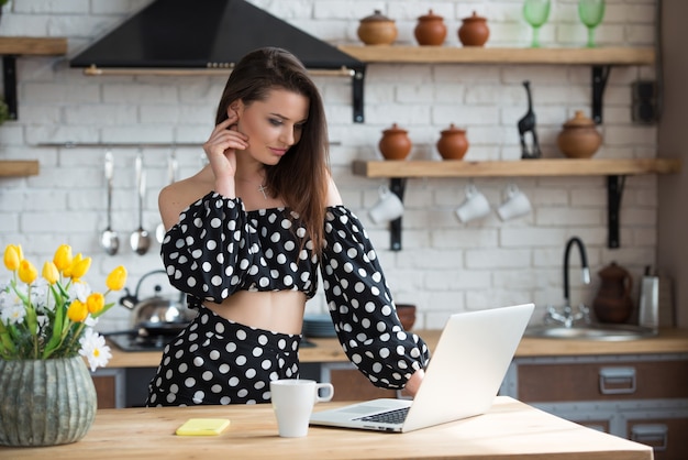 Blogger atractiva chica morena con un vestido de lunares sentado en la acogedora cocina y leyendo noticias en la computadora portátil en la mesa de madera.