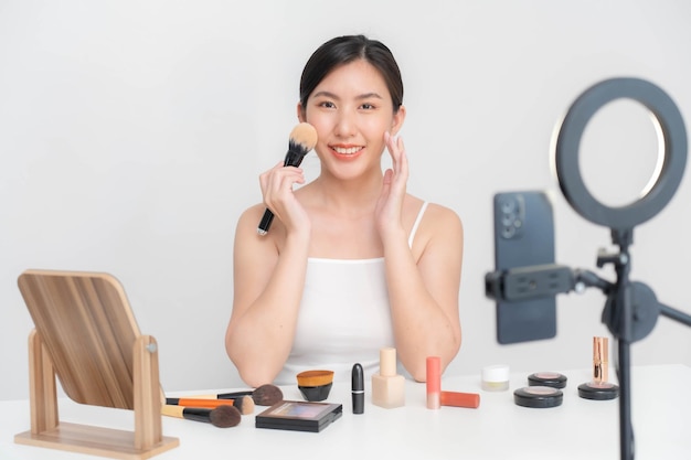 Una blogger de belleza asiática usa un pincel de rubor para cepillarse la cara durante un tutorial de maquillaje en línea en vivo a través de Internet