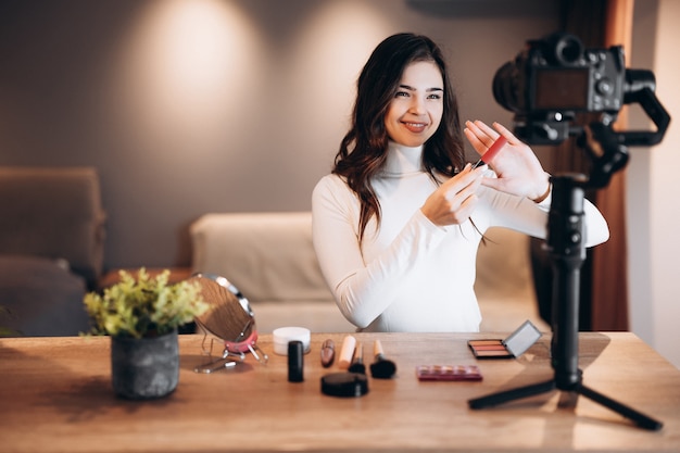 Blogger de belleza bastante femenina filmando un tutorial de rutina de maquillaje diario en la cámara. Mujer joven influyente revisión de productos cosméticos de transmisión en vivo en estudio casero. Trabajo de vlogger. Bricolaje.