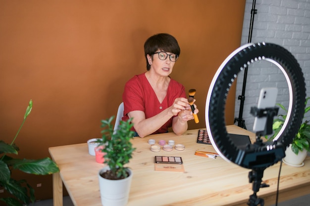 Blogger de belleza femenina de mediana edad o influencer con producto de maquillaje grabando video en la habitación en casa u